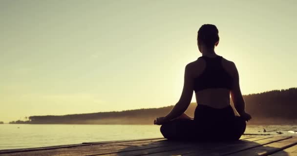 スリムな女の子は、日没時に川の近くの桟橋に座ってロータスポーズで瞑想しています. — ストック動画