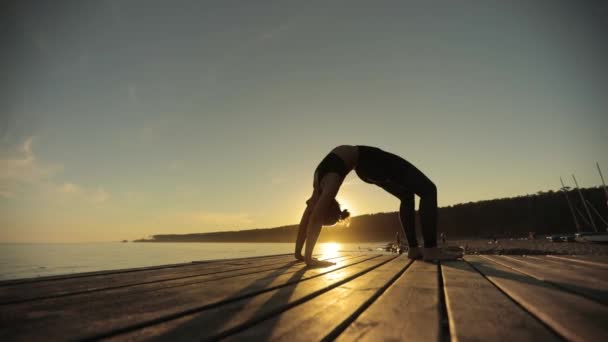 Gün batımında nehir iskelesinde Wheel Pose yoga ince kız silueti. — Stok video