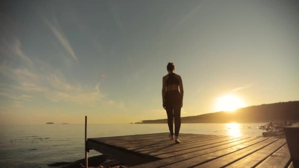 Menina desportiva está praticando ioga em pose de árvore em pé no cais pelo rio ao pôr do sol . — Vídeo de Stock