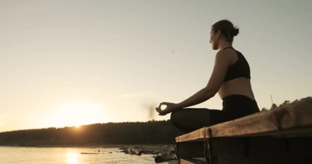 Молода мама роздумує, сидячи на річковому причалі на заході сонця в позі лотоса . — стокове відео