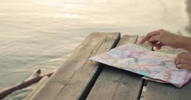 Woman chooses the route of her trip at map on sea pier at sunset, hands closeup. — Stock Video