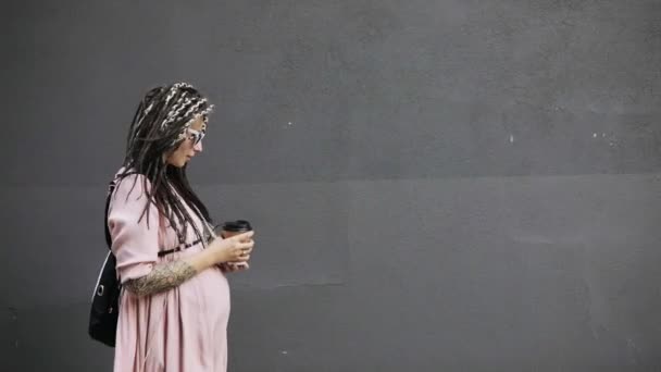 Gelukkige jonge zwangere vrouw hipster wandelen in de straat in de stad met koffie. — Stockvideo