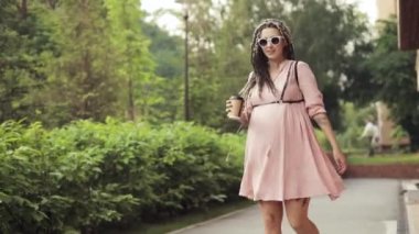 Gülümseyen hamile genç kadın şehir parkında yürüyor ve kameraya bakıyor.