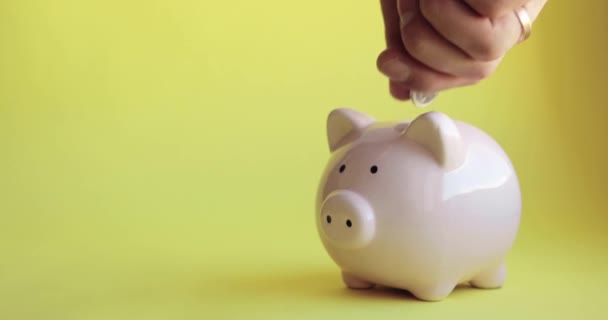 Mans hand putting coins in piggy bank of pig shape on yellow background. — Stock Video