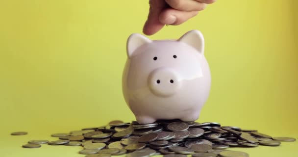 Man putting coins in piggy bank standing in pile of coins, hand closeup. — Stock Video