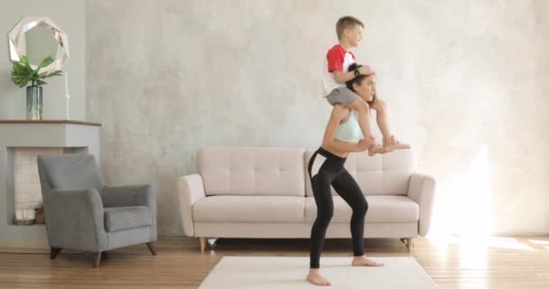 Vrouw maakt hurken met haar zoon zittend op haar schouders, thuis aan het trainen. — Stockvideo