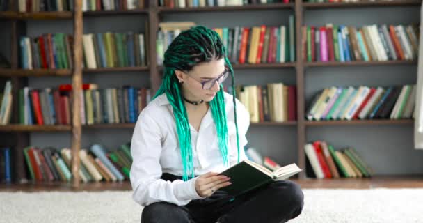 Молодая студентка с зелеными волосами в очках читает книгу в библиотеке . — стоковое видео