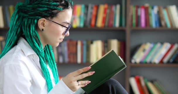 Retrato de menina estudante com dreadlocks verdes em óculos lê livro na biblioteca . — Vídeo de Stock