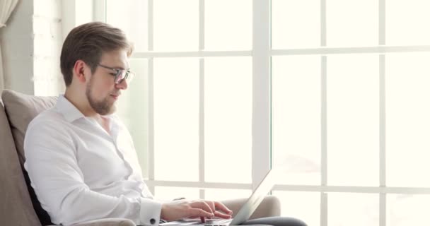 Ο άνθρωπος με τα γυαλιά εργάζεται δακτυλογράφηση σε φορητό υπολογιστή στο γραφείο κοντά στο παράθυρο, πλευρική άποψη. — Αρχείο Βίντεο