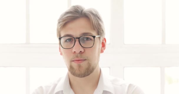 Retrato de hombre joven en gafas y camisa blanca cerca de la ventana mirando a la cámara . — Vídeos de Stock