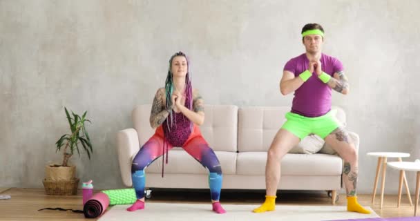 Funny man and woman in colourful sportswear doing squats exercises at home. — Stock Video