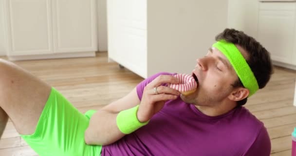 Mężczyzna lubi jeść pączki leżące na macie fitness w odzieży sportowej w domu. — Wideo stockowe