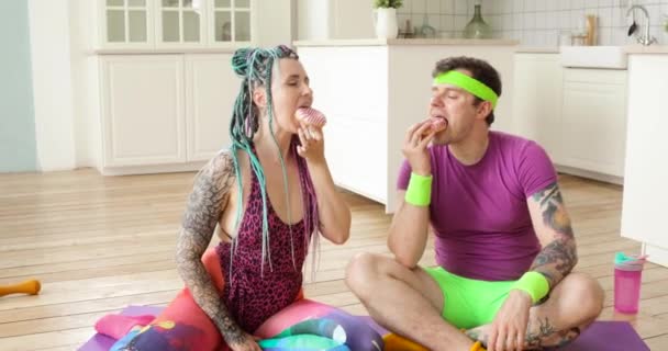 Copule hombre y mujer disfrutando de comer donas sentados en la colchoneta de fitness en casa . — Vídeos de Stock