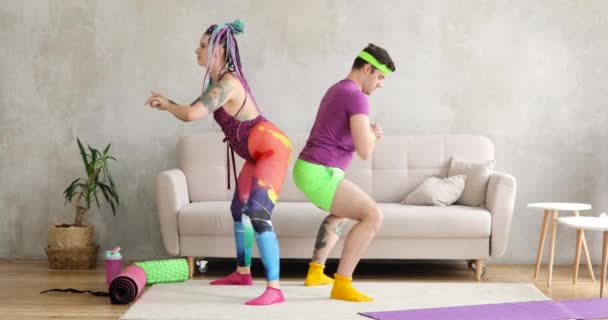 ผู้ชายและผู้หญิงตลกในชุดกีฬาที่มีสีสัน ทําการออกกําลังกาย squats ที่บ้าน . — วีดีโอสต็อก