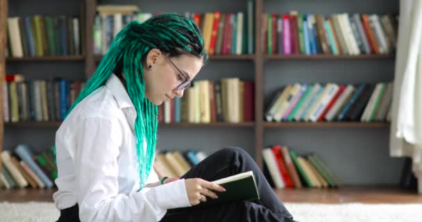 Étudiante avec dreadlocks verts dans des lunettes lit livre dans la bibliothèque, vue de côté. — Video