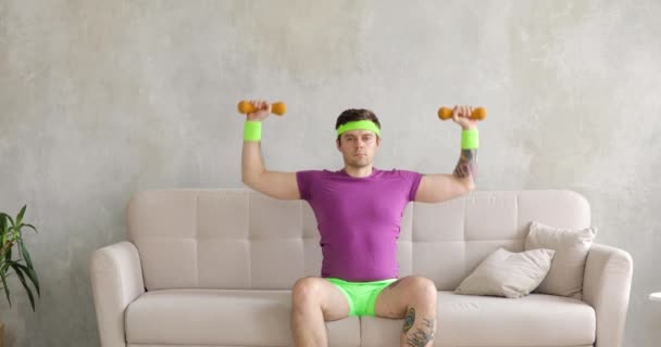 Fjollet sjov mand narcissist løfte håndvægte ved hænder sidder på sofaen derhjemme. – Stock-video