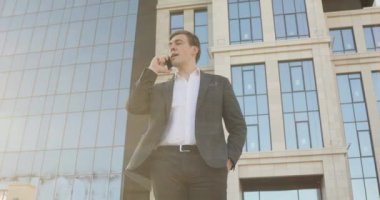 İş adamı şehir merkezindeki gökdelenin yanındaki akıllı telefondan konuşuyor..