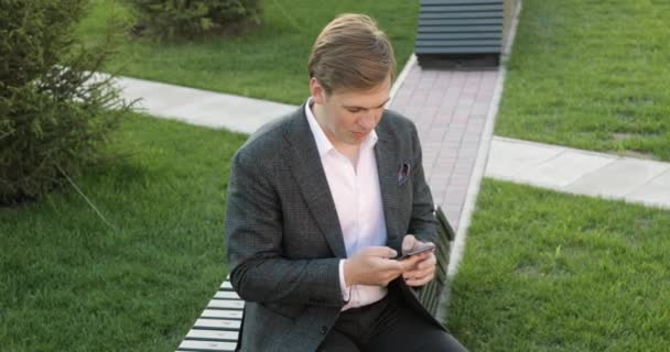 Podnikatel je psaní na smartphone procházení on-line sedí na lavičce v parku. — Stock video
