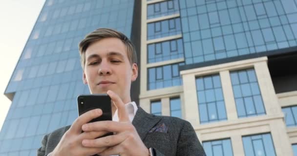 Молодой бизнесмен печатает на смартфоне возле офисного здания в центре города . — стоковое видео