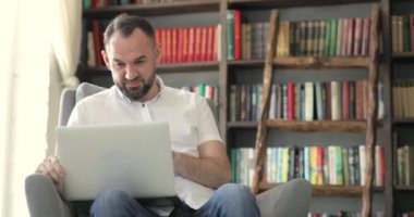 Yetişkin bir adam konuşuyor. Arkasında kitap raflarında oturmuş video görüşmesi yapıyor..