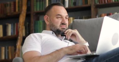 Yetişkin bir adamın portresi ev ofisinin koltuğunda dizüstü bilgisayarla çalışıyor..