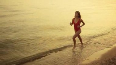 Kırmızı mayo giymiş genç ve çekici bir kadın gün batımında kumlu sahilde koşuyor..