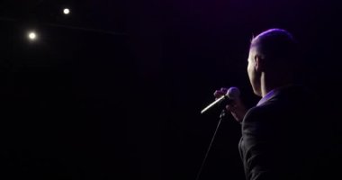 Adam ayağa kalkar, komedyen gibi mikrofon şakaları yapar sahnede, arka planda.