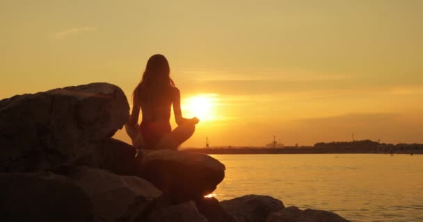 Femme médite assis sur des pierres sur la plage de la mer au coucher du soleil dans la ville, vue arrière. — Video