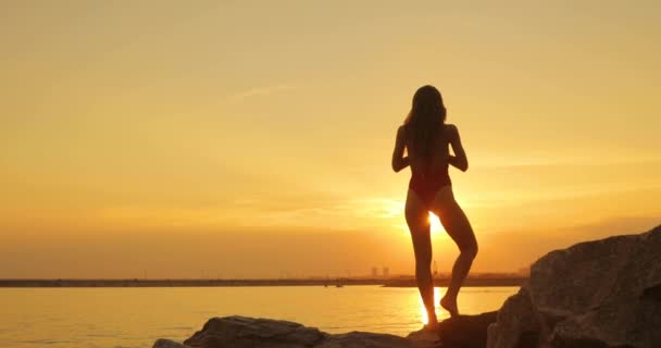 Mujer en traje de baño está haciendo pose de árbol practicando yoga en la playa al atardecer . — Vídeos de Stock