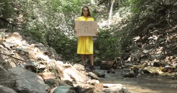 Kobieta stoi w pobliżu strumienia ścieków w lesie z plakatem uratować planetę. — Wideo stockowe