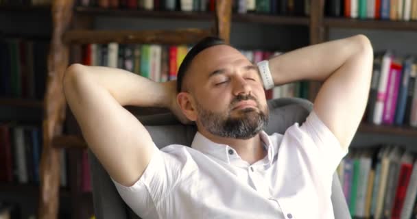 Dojrzały mężczyzna marzy o spoczywaniu w fotelu w bibliotece z rękami za głową. — Wideo stockowe