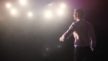 Adam ayağa kalkıp komedyen şakaları yapıyor mikrofon kulaklığıyla sahnede, arka planda.