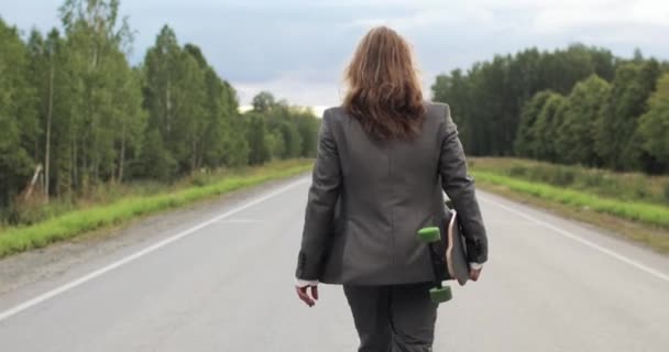 オフィススーツの男でスケートボードを手に街の外の道路を歩いて. — ストック動画