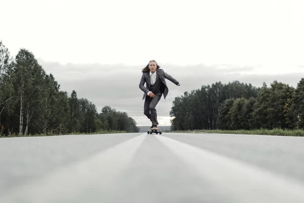 Homem em terno de escritório está montando skate longboard estrada abaixo fora da cidade . — Fotografia de Stock