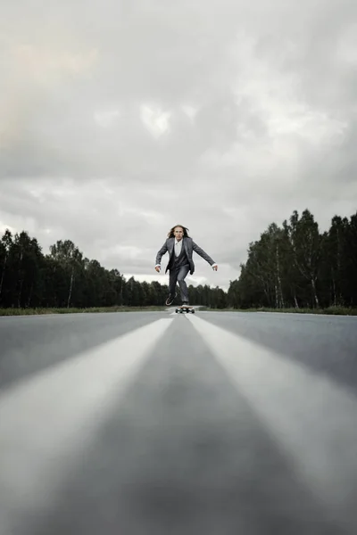 Handsome man in office suit with longboard walking down road in city outskirts.