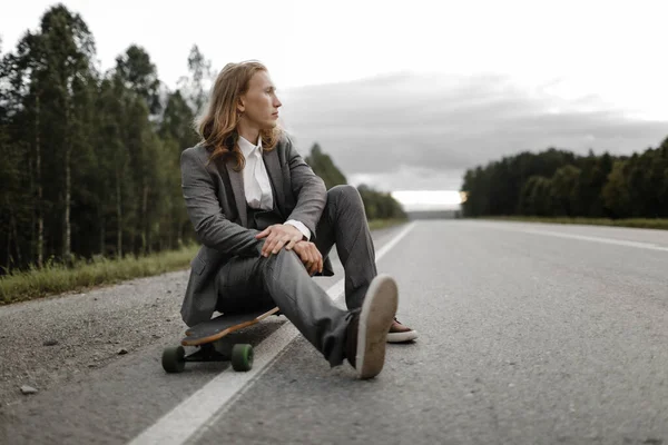 Snygg man i kontorskostym med longboard promenader längs vägen i utkanten av staden. — Stockfoto