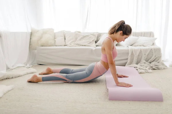 Joven chica de deportes en ropa deportiva está haciendo yoga y estiramiento en casa en una habitación luminosa. — Foto de Stock