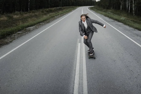 Άντρας με κοστούμι γραφείου ιππεύει skateboard longboard κάτω από το δρόμο έξω από την πόλη. Εικόνα Αρχείου