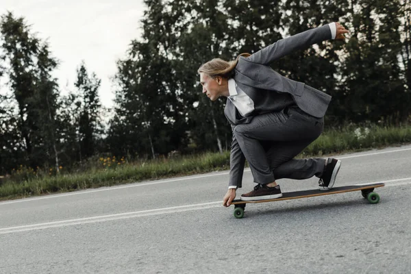 정장을 입은 남자가 도시 외곽 도로에서 스케이트보드를 타고 있습니다. 로열티 프리 스톡 사진