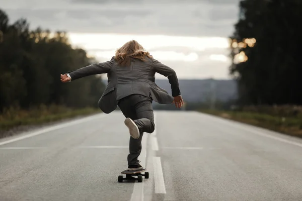 Άντρας με κοστούμι γραφείου ιππεύει skateboard longboard κάτω από το δρόμο έξω από την πόλη. Φωτογραφία Αρχείου