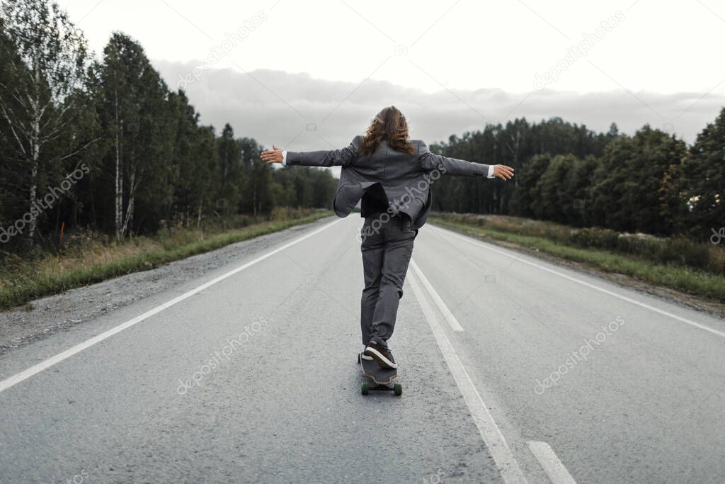Man in office suit is riding skateboard longboard down road outside the city.