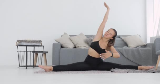 Sporty jeune femme fait des exercices d'étirement assis sur le tapis appuyé sur les jambes. — Video