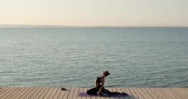 Güvercin pozisyonunda yoga yapan kadın nehir ahşap iskelede spor minderinde oturuyor..