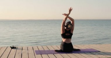 Yoga yapan kadın nehir rıhtımında mindere oturup yan yatarak esneme hareketleri yapıyor..
