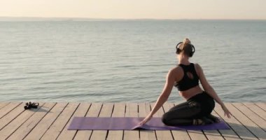 Yoga yapan bir kadın iskelede dizlerinin üzerine çöküp kendini paspaslıyor..