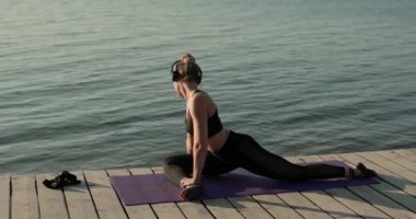 Kadın yoga yapıyor bacaklarını esnetiyor ve nehir rıhtımında mindere yaslanıyor..