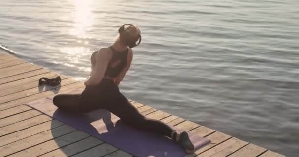 Žena cvičí jógu dělá holubí pózu a naklání se dopředu na podložku na říčním molu. — Stock video