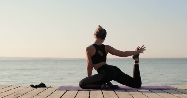 Wanita berlatih yoga melakukan pose merpati peregangan kakinya pada tikar di dermaga sungai. — Stok Video