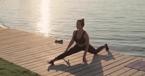 Sportovní mladá žena dělá podélné rozdělení sedí na molu u řeky. — Stock video