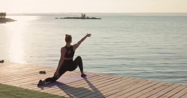 Mujer está practicando yoga haciendo salto guerrero yoga pose en el muelle del río, vista trasera. — Vídeos de Stock